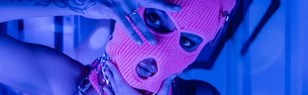 Provocante donna in passamontagna posa con le mani vicino al viso a parete con graffiti in luce blu al neon, banner — Foto stock