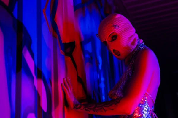 Женщина в балаклаве прикасается к голубой стене граффити при взгляде на камеру при красном освещении — стоковое фото