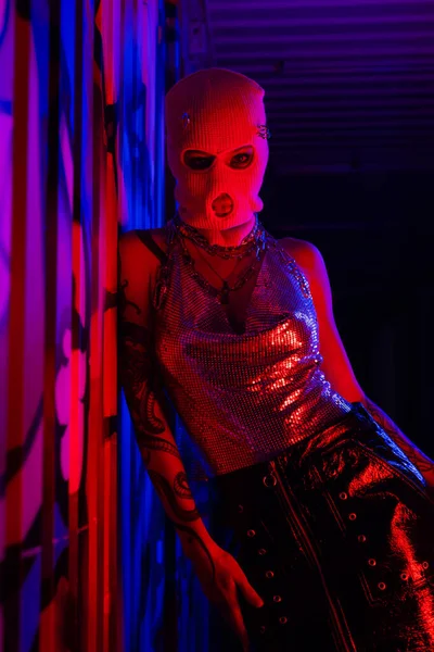 Anonyme tätowierte Frau mit Sturmhaube und sexy Outfit lehnt an Wand mit Graffiti in blauem und rotem Licht — Stockfoto
