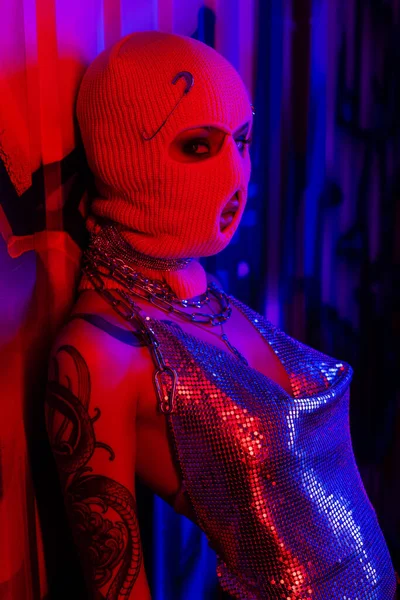Сексуальная женщина с татуировками в вязаной балаклаве и серебряном топе, смотрящая на камеру рядом с граффити на синем фоне при красном освещении — стоковое фото