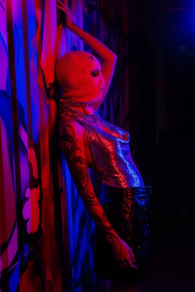 Side view of tattooed woman in silver top and balaclava posing in blue and red light near wall with graffiti — Stock Photo