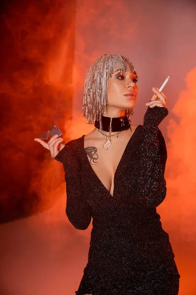 Mujer tatuada extravagante en peluca de plata y vestido sexy posando con cigarrillo sobre fondo rojo y naranja con humo - foto de stock