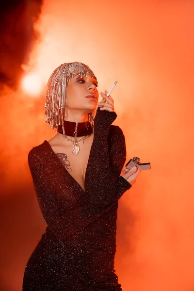 Сексуальная женщина с татуировками в парике из страз и черном платье из люрекса, стоящее с сигаретой на оранжевом фоне с дымом — стоковое фото