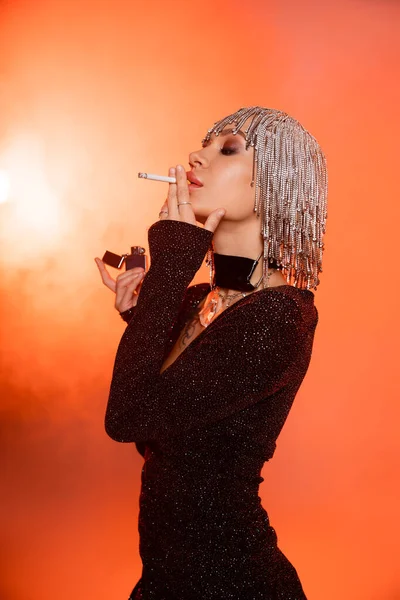 Sexy model in black lurex dress and jewelry headwear standing with cigarette on orange background with smoke — Stock Photo