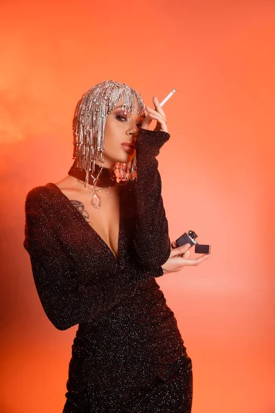 Donna glamour in parrucca argentata e abito lurex nero con in mano sigaretta e accendino su sfondo rosa e arancione con fumo — Foto stock