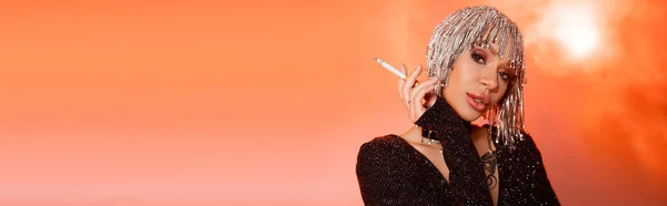 Porträt einer sinnlich tätowierten Frau in metallischer Kopfbedeckung mit Zigarette auf korallenrosa rauchigem Hintergrund, Banner — Stockfoto