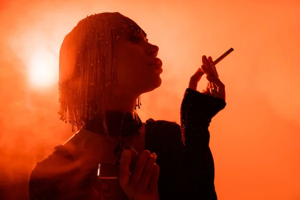 Гламурная женщина в металлическом парике позирует с сигаретой на оранжевом фоне с задним светом и дымом — стоковое фото