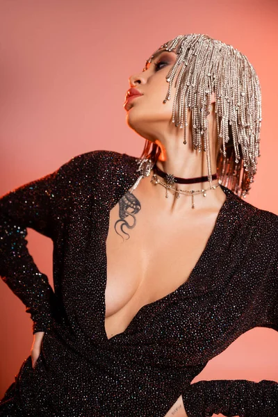 Mulher tatuada com busto sexy posando em peruca de jóias e vestido lurex preto enquanto olha para o fundo rosa coral — Fotografia de Stock