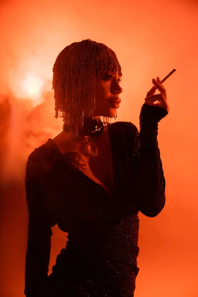 Femme sexy en casque métallique et robe noire debout avec cigarette sur fond orange avec de la fumée — Photo de stock