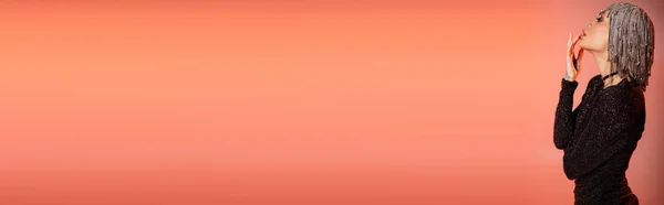 Vista laterale della donna glamour in copricapo metallico e abito lurex nero toccare le labbra su sfondo rosa corallo, banner — Foto stock