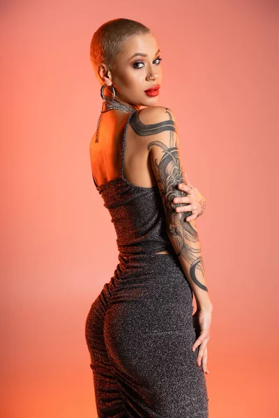 Tattooed woman with makeup and red lips looking at camera while posing in sexy outfit on coral pink background — Stock Photo