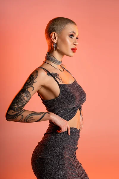 Tattooed woman in grey lurex skirt and crop top looking away on coral pink background — Stock Photo