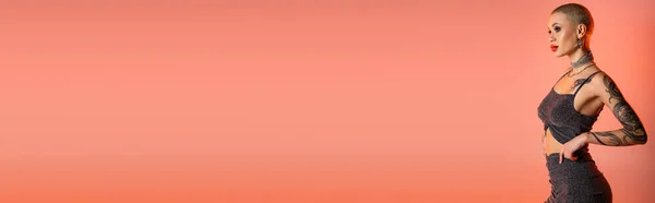 Femme tatouée aux cheveux courts en jupe et haut de culture debout et regardant loin sur fond rose corail, bannière — Photo de stock
