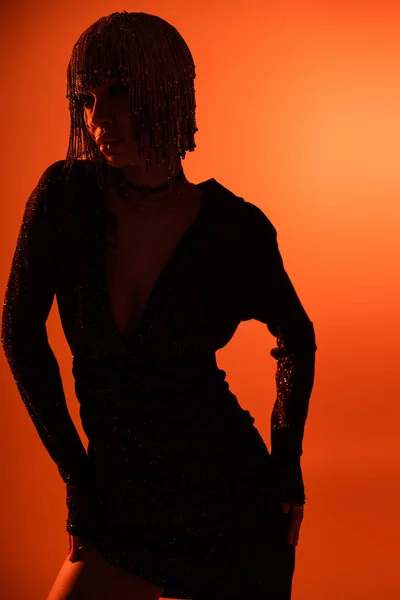 Silueta oscura de mujer joven glamour en peluca metálica y vestido sexy negro posando con las manos en las caderas sobre fondo naranja - foto de stock