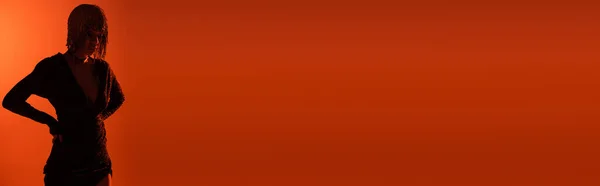 Затененный силуэт сексуальной женщины, стоящей с руками на талии на красном и оранжевом фоне с копировальным пространством, баннером — стоковое фото