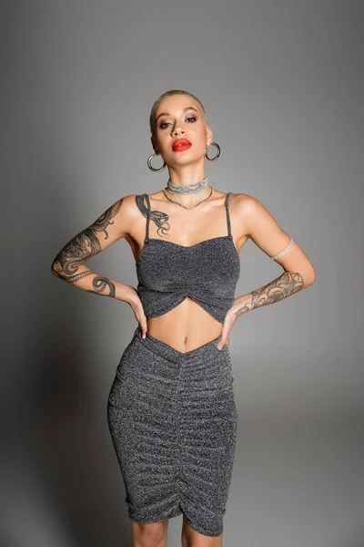 Mujer tatuada extravagante con pelo corto y maquillaje brillante mirando a la cámara mientras posa con las manos en la cintura sobre fondo gris - foto de stock