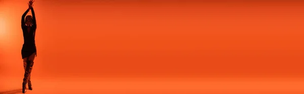Повна довжина молодої жінки в сексуальній сукні і довгі чоботи позує з піднятими руками на помаранчевому фоні, банер — стокове фото