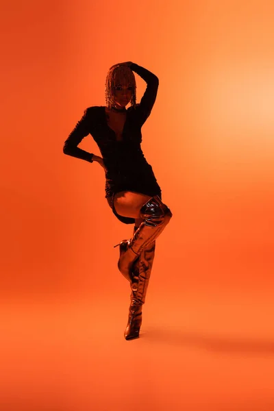Повна довжина сексуальної моделі в модних чоботях і чорна сукня позує на одній нозі на помаранчевому фоні — стокове фото