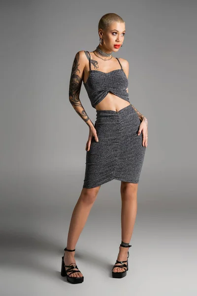 Full length of sexy tattooed woman in lurex skirt and crop top standing with hands on hips and looking away on grey background — Stock Photo