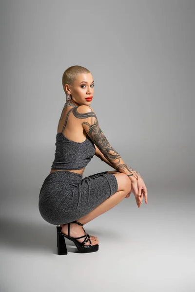 Full length of sexy tattooed woman in lurex skirt and heeled sandals sitting on haunches on grey background — Stock Photo