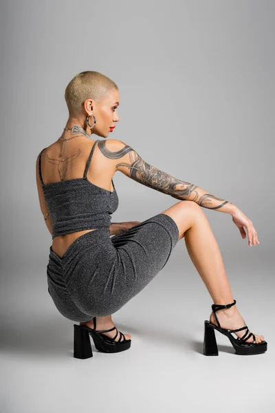 Full length of glamour tattooed woman in sexy outfit posing on haunches and looking away on grey background — Stock Photo