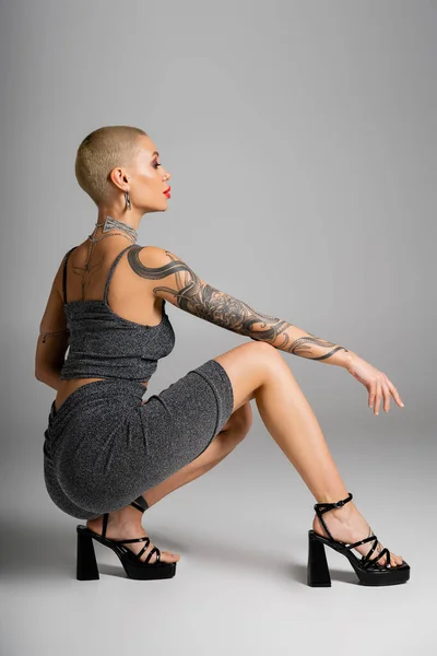 Vista lateral de mujer glamurosa tatuada en traje sexy sentada sobre horquillas sobre fondo gris - foto de stock
