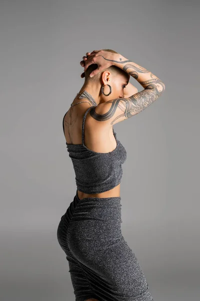 Junge Frau mit sexy tätowierten Körper posiert in stylischer Lurex-Kleidung mit den Händen auf dem Kopf isoliert auf grau — Stockfoto