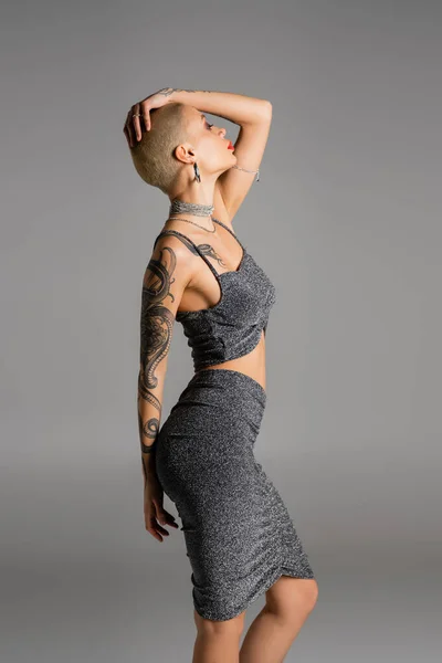 Side view of sexy tattooed woman in stylish lurex clothes posing with hand above head isolated on grey — Stock Photo