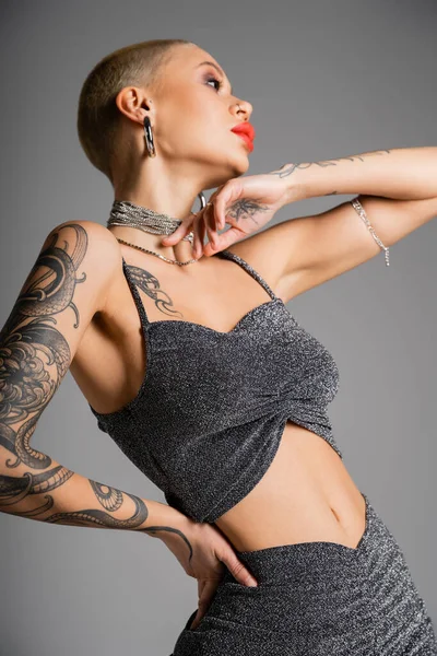 Low angle view of tattooed woman in stylish crop top and necklaces posing with hand on hip isolated on grey — Stock Photo