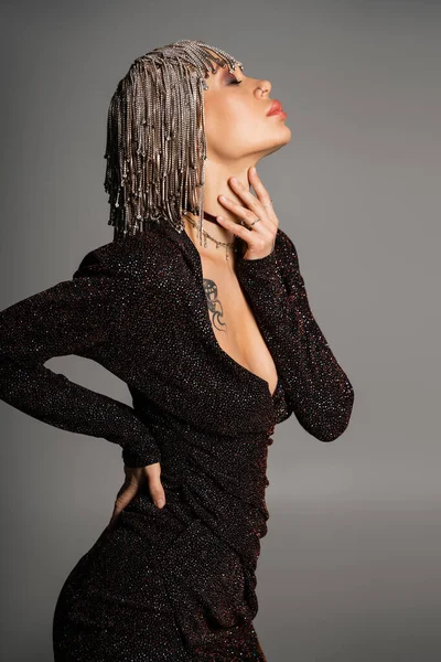 Sensuale donna in abito sexy e gioielli parrucca toccante collo mentre posa con mano sul fianco isolato su grigio — Foto stock
