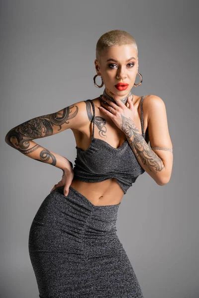 Seducente donna tatuata in lurex gonna e crop top toccare collane mentre posa con mano sul fianco isolato su grigio — Foto stock