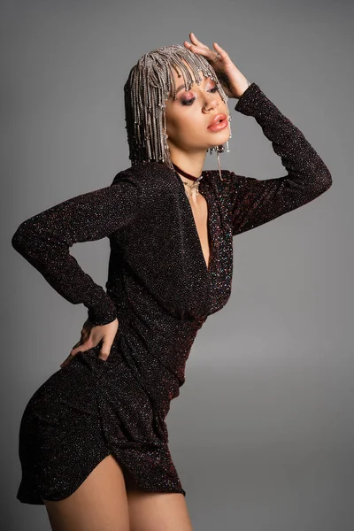 Extravagante Frau in schwarzem sexy Kleid, die silberne Perücke berührt, während sie mit der Hand an der Hüfte isoliert auf grau steht — Stockfoto