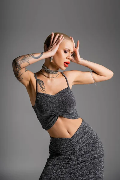 Экстравагантная татуированная женщина в сексуальном наряде, держась за руки рядом с головой, изолированной на серой — стоковое фото