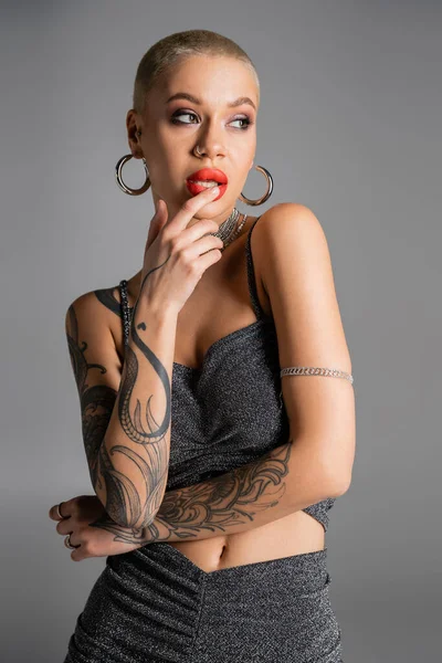 Соблазнительная татуированная женщина в серьгах с обручем и топе из люрекса касается красных губ и смотрит в сторону изолированной на сером — стоковое фото