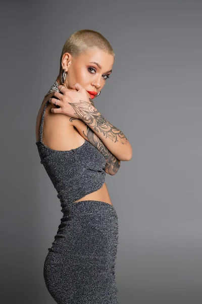 Mujer tatuada de pelo corto con ropa glamurosa abrazándose mientras mira a la cámara aislada en gris - foto de stock