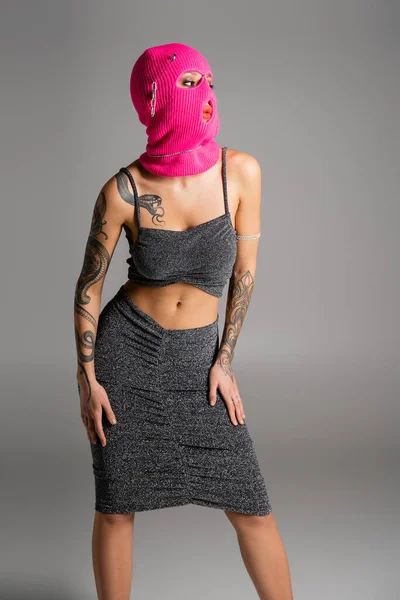 Verführerische tätowierte Frau in Lurex-Kleidung und rosa Sturmhaube, die isoliert auf grau schaut — Stockfoto