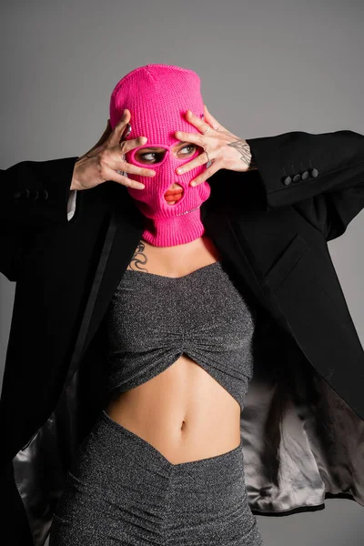 Сексуальная женщина с татуировками в стильной одежде, держась за руки рядом с лицом в розовой балаклаве, глядя в сторону изолированной на сером — стоковое фото