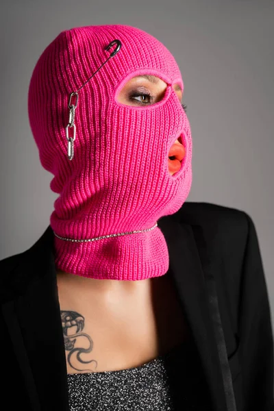 Porträt einer extravaganten Frau in schwarzem Blazer und pinkfarbener Sturmhaube, die isoliert auf grau blickt — Stockfoto