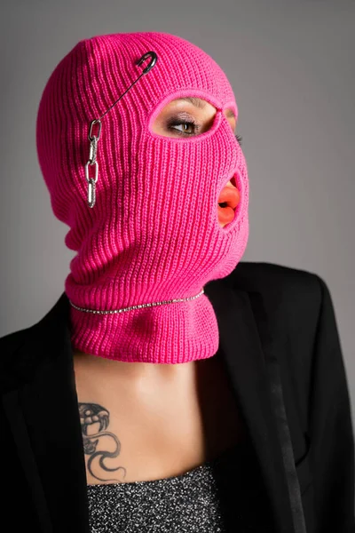 Retrato de mulher extravagante em blazer preto e balaclava rosa olhando para longe isolado em cinza — Fotografia de Stock