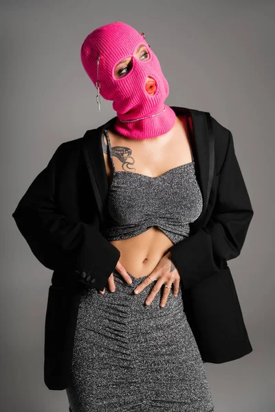 Сексуальная женщина с татуировками, позирующая в модной одежде и розовой балаклаве изолированная на сером — стоковое фото