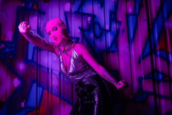 Mulher sedutora no topo metálico e balaclava segurando corrente de prata perto da parede com grafite colorido em luz roxa — Fotografia de Stock