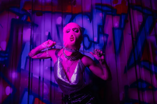 Leidenschaftliche tätowierte Frau in rosa Sturmhaube und metallischem Oberteil posiert mit Kette in Wandnähe mit Graffiti in lila Beleuchtung — Stockfoto