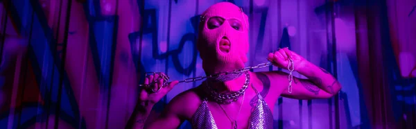 Страстная женщина в балаклаве позирует с серебряной цепью рядом с граффити в фиолетовом свете, баннер — стоковое фото