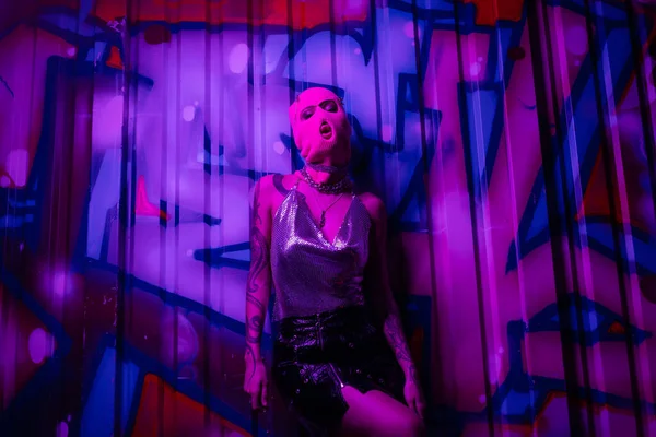 Donna sexy in passamontagna e top in argento con gonna nera in posa vicino graffiti colorati in luce viola — Foto stock