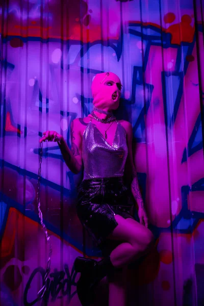 Provocative woman in pink balaclava and shiny top standing with silver chain near colorful graffiti in purple neon light — Stock Photo