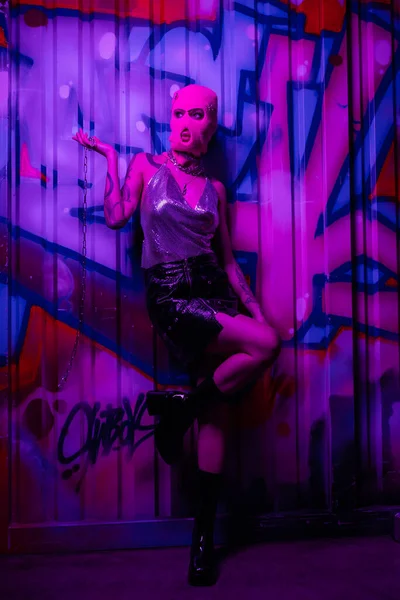 Full length of stylish woman in balaclava holding silver chain and looking away near wall with graffiti in purple light — Stock Photo