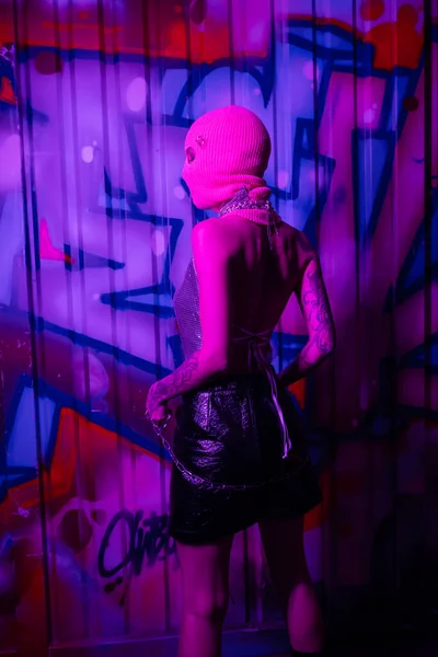Задний вид сексуальной женщины в балаклаве и черной кожаной юбке, стоящей с серебряной цепью рядом с красочными граффити в фиолетовом неоновом свете — стоковое фото