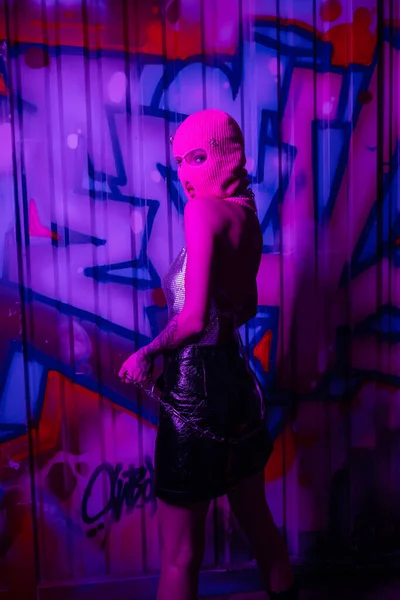 Seductive woman in pink balaclava posing with silver chain and looking at camera near wall with graffiti in purple lighting — Stock Photo