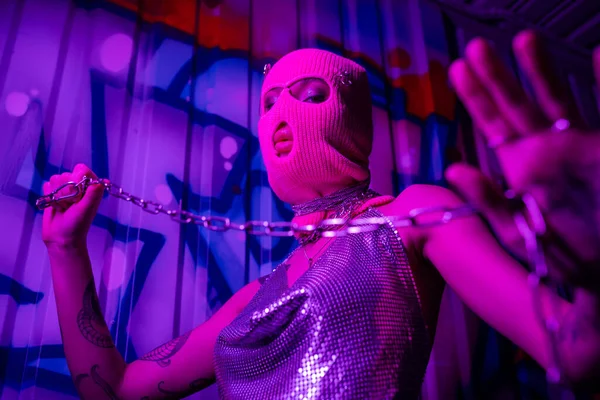Vue à angle bas de femme sexy en cagoule rose et haut brillant tenant chaîne d'argent et regardant la caméra en lumière au néon violet — Photo de stock