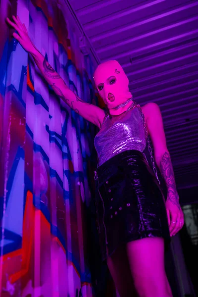 Low angle view of hot woman in black leather skirt and balaclava looking at camera near colorful graffiti in purple lighting — Stock Photo
